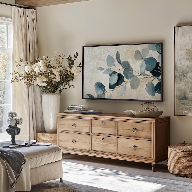 May include: A light wood dresser with six drawers and brass knobs. The dresser is in a living room with a large TV above it, a vase of white flowers, and a throw blanket on a light-colored ottoman.