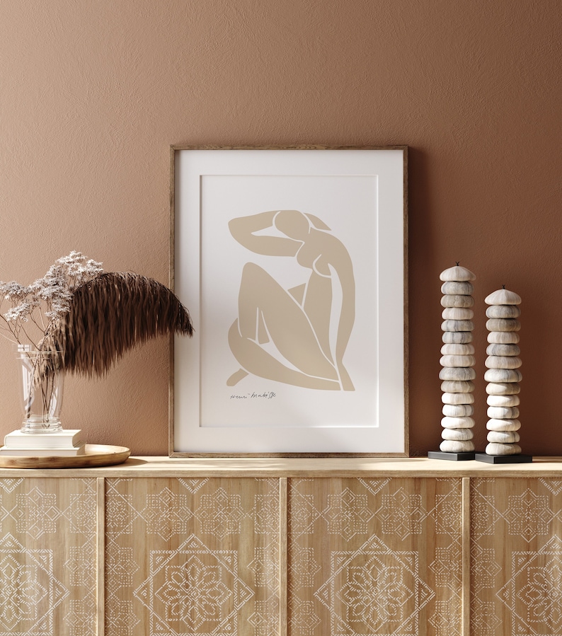 May include: A framed print of a minimalist line drawing of a nude figure in a seated pose. The print is on a white background with a brown wooden frame. The print is hanging on a wall with a light brown background.