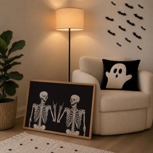 May include: A rectangular framed print featuring two white skeletons on a black background, with one skeleton making a rock-and-roll hand gesture. The print is placed on the floor next to a cream-colored armchair with a black ghost-patterned pillow.