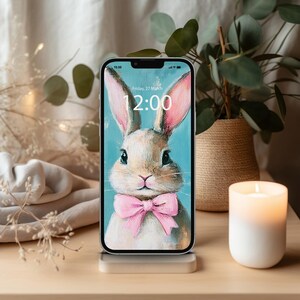 May include: A smartphone displays a bunny illustration with a pink bow tie and the time 12:00. The phone is on a clear stand, with a lit candle and a plant in the background. The screen shows the date, Friday, March 27.