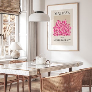 May include: A framed art print featuring a Matisse design with pink floral elements on a beige background. The print hangs above a dining table with woven chairs. A white pendant light hangs above the table, and a window with a bamboo blind is visible.