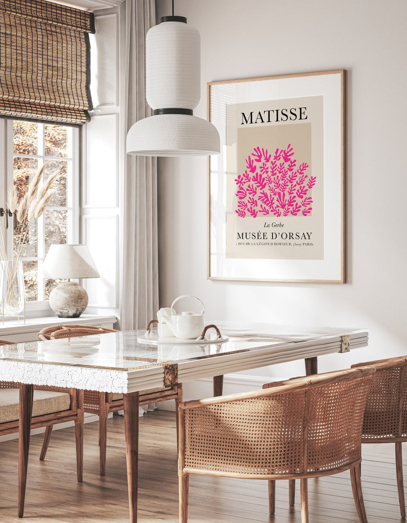 May include: A framed art print featuring a Matisse design with pink floral elements on a beige background. The print hangs above a dining table with woven chairs. A white pendant light hangs above the table, and a window with a bamboo blind is visible.