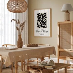 K&ouml;nnte beinhalten: Ein beiger Essbereich mit einem Holztisch und St&uuml;hlen, einer H&auml;ngeleuchte aus Weidengeflecht, einem gerahmten Schwarz-Wei&szlig;-Abstrakt-Kunstdruck mit dem Text "Henri Matisse" und einem Schrank aus Holz mit einer Front aus Weidengeflecht.