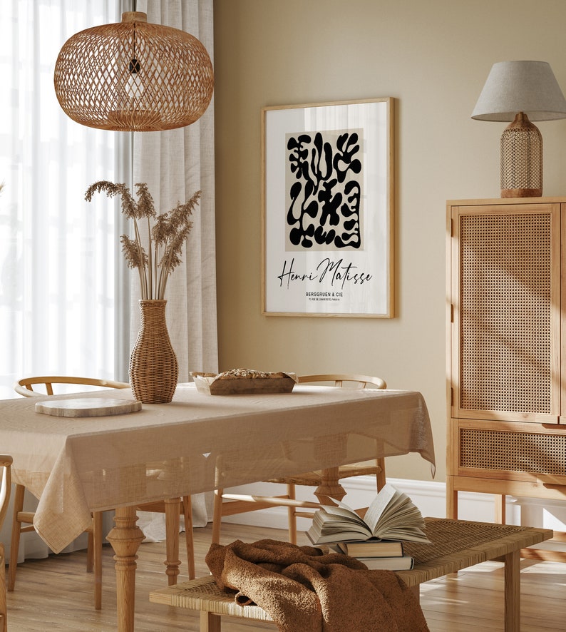 K&ouml;nnte beinhalten: Ein beiger Essbereich mit einem Holztisch und St&uuml;hlen, einer H&auml;ngeleuchte aus Weidengeflecht, einem gerahmten Schwarz-Wei&szlig;-Abstrakt-Kunstdruck mit dem Text "Henri Matisse" und einem Schrank aus Holz mit einer Front aus Weidengeflecht.