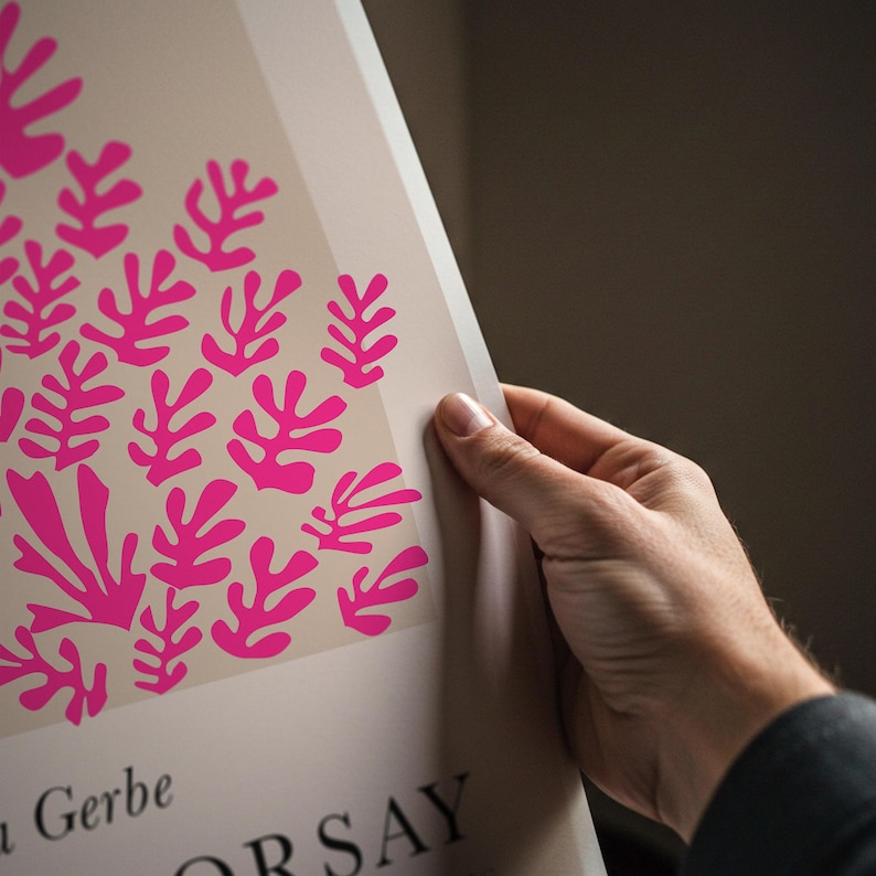 May include: A poster featuring a pink floral design on a cream background. The artwork is held by a hand, showcasing the edge of the print. The poster has text at the bottom, with the name "Gerbe" visible.