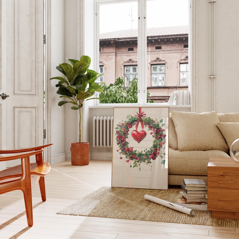 May include: A framed print of a red heart hanging from a green wreath with red berries. The wreath is surrounded by green leaves and vines. The print is hanging on a white wall in a living room setting.