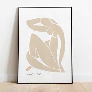 May include: A beige abstract line drawing of a person sitting with their arm raised above their head. The drawing is framed in a black frame and the text "femme - made by me" is at the bottom of the drawing.