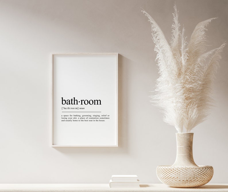 May include: A black and white printable wall art print with the word "bath-room" defined as a space for bathing, grooming, singing, relief or losing your shirt, a place of contention sometimes and usually home to the best seat in the house. The print is framed in a white frame and is hanging on a white wall. A white pampas grass bouquet is in a woven vase on a white shelf.