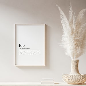 May include: A white framed print with the word "loo" defined as a space for bathing, grooming, singing, relief or losing your shirt, a place of contention sometimes and usually home to the best seat in the house. The print is displayed on a white wall with a white vase with a white pampas grass arrangement.