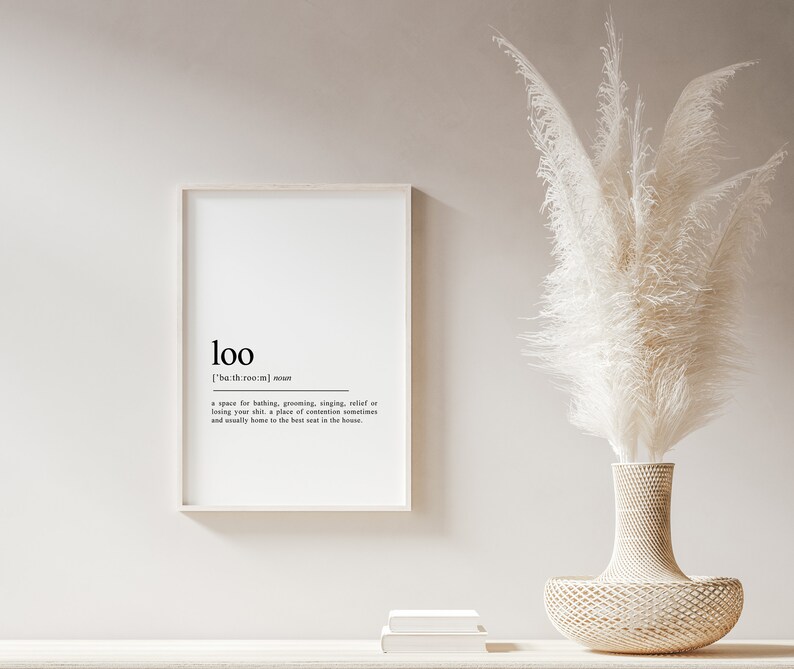 May include: A white framed print with the word "loo" defined as a space for bathing, grooming, singing, relief or losing your shirt, a place of contention sometimes and usually home to the best seat in the house. The print is displayed on a white wall with a white vase with a white pampas grass arrangement.