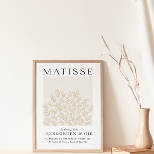 May include: A framed print of a beige abstract art piece by Henri Matisse titled "La Gerbe" (1953). The print features a cluster of stylized beige leaves against a white background. The artist's name, "MATISSE", is printed in black at the top of the print. The print is framed in a light wood frame. The text below the art piece reads "La Gerbe (1953) BERGGRUEN & CIE 70, RUE DE L'UNIVERSIT&Eacute;, PARIS-VII Du Vendredi 27 F&eacute;vrier au Samedi 28 Mars 1953."