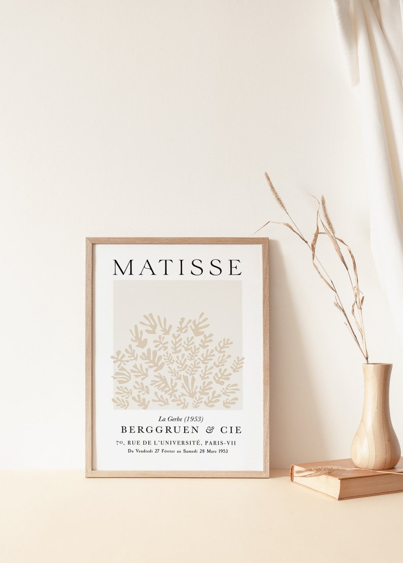 May include: A framed print of a beige abstract art piece by Henri Matisse titled "La Gerbe" (1953). The print features a cluster of stylized beige leaves against a white background. The artist's name, "MATISSE", is printed in black at the top of the print. The print is framed in a light wood frame. The text below the art piece reads "La Gerbe (1953) BERGGRUEN & CIE 70, RUE DE L'UNIVERSIT&Eacute;, PARIS-VII Du Vendredi 27 F&eacute;vrier au Samedi 28 Mars 1953."