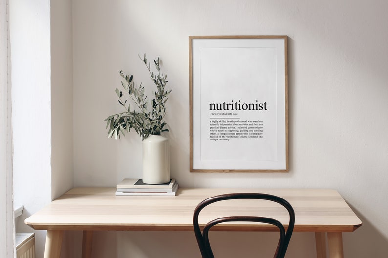 May include: A framed print with the word "nutritionist" and its definition. The print is hanging on a white wall above a wooden desk with a vase of green branches and a stack of books.