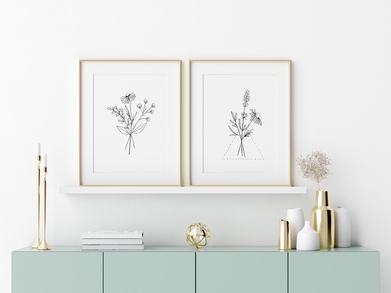 May include: Two framed black and white line drawings of floral bouquets, one with a triangle shape, on a white wall above a light blue cabinet with gold accents.