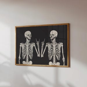 May include: A rectangular wall art print in a wooden frame featuring two off-white skeletons against a solid black background. Both skeletons are depicted from the waist up, with one skeleton making a rock-and-roll hand gesture with its fingers.