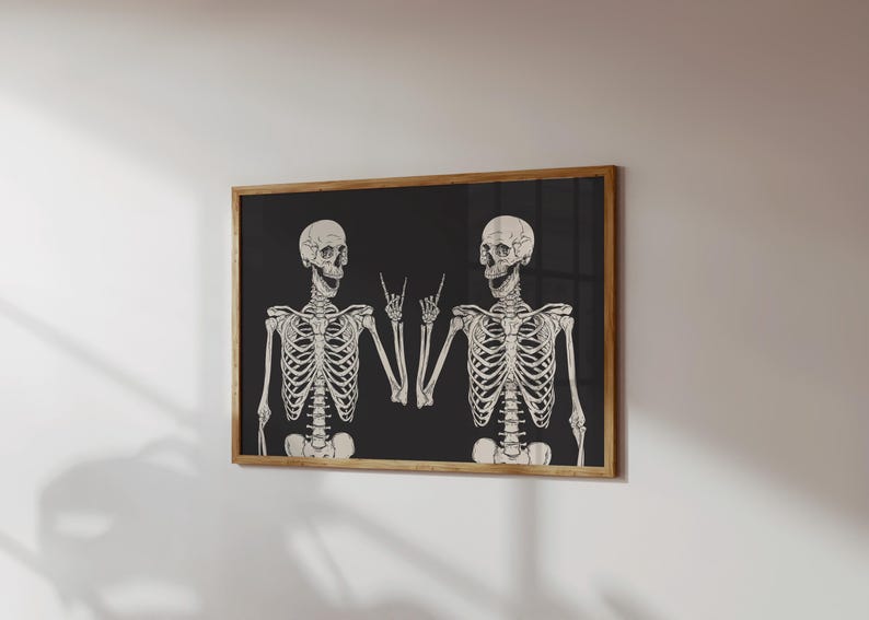 May include: A rectangular wall art print in a wooden frame featuring two off-white skeletons against a solid black background. Both skeletons are depicted from the waist up, with one skeleton making a rock-and-roll hand gesture with its fingers.