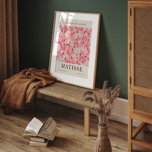May include: A framed print of a Matisse artwork titled "Papiers Decoupes" featuring pink and white cut paper flowers on a light pink background. The print is hanging on a green wall above a wooden bench with a brown blanket draped over it. A wicker vase with dried flowers sits on the floor.