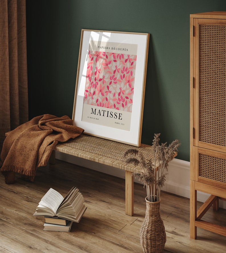May include: A framed print of a Matisse artwork titled "Papiers Decoupes" featuring pink and white cut paper flowers on a light pink background. The print is hanging on a green wall above a wooden bench with a brown blanket draped over it. A wicker vase with dried flowers sits on the floor.