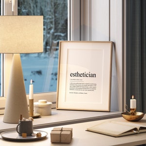 May include: A framed print with the word "esthetician" and its definition. The print is on a white background with black text. The frame is light brown wood. The print is sitting on a white desk with a brown lamp, two candles, a small white container, a brown mug, a small brown box, and a book.
