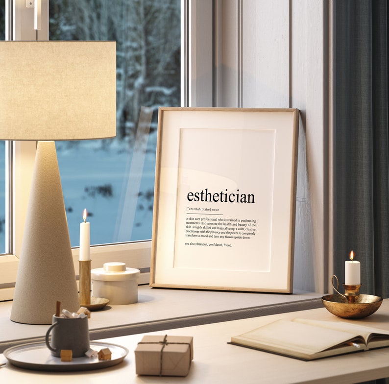 May include: A framed print with the word "esthetician" and its definition. The print is on a white background with black text. The frame is light brown wood. The print is sitting on a white desk with a brown lamp, two candles, a small white container, a brown mug, a small brown box, and a book.