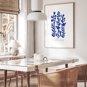 May include: A white dining table with a marble top and a white ceramic teapot on a white plate. Two wicker chairs are placed on either side of the table. A white pendant light hangs above the table. A framed print with blue botanical illustrations and the text "LE JARDIN BOTANIQUE" is on the wall behind the table.