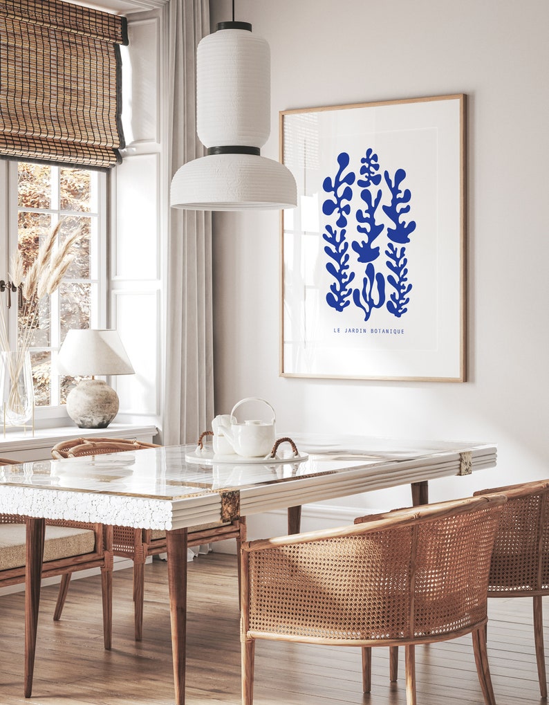 May include: A white dining table with a marble top and a white ceramic teapot on a white plate. Two wicker chairs are placed on either side of the table. A white pendant light hangs above the table. A framed print with blue botanical illustrations and the text "LE JARDIN BOTANIQUE" is on the wall behind the table.