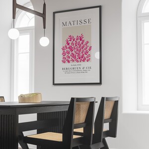 May include: A black framed poster with the text "MATISSE" in white letters. The poster features a pink abstract design of leaves and branches on a beige background. The text "La Gerbe (1953)" is in black letters below the design. The text "BERGGRUEN & CIE" is in black letters below the date. The text "70, RUE DE L'UNIVERSIT&Eacute;, PARIS-VII" is in black letters below the company name. The text "Du Samedi 27 Fevrier au Samedi 28 Mars 1953" is in black letters below the address.