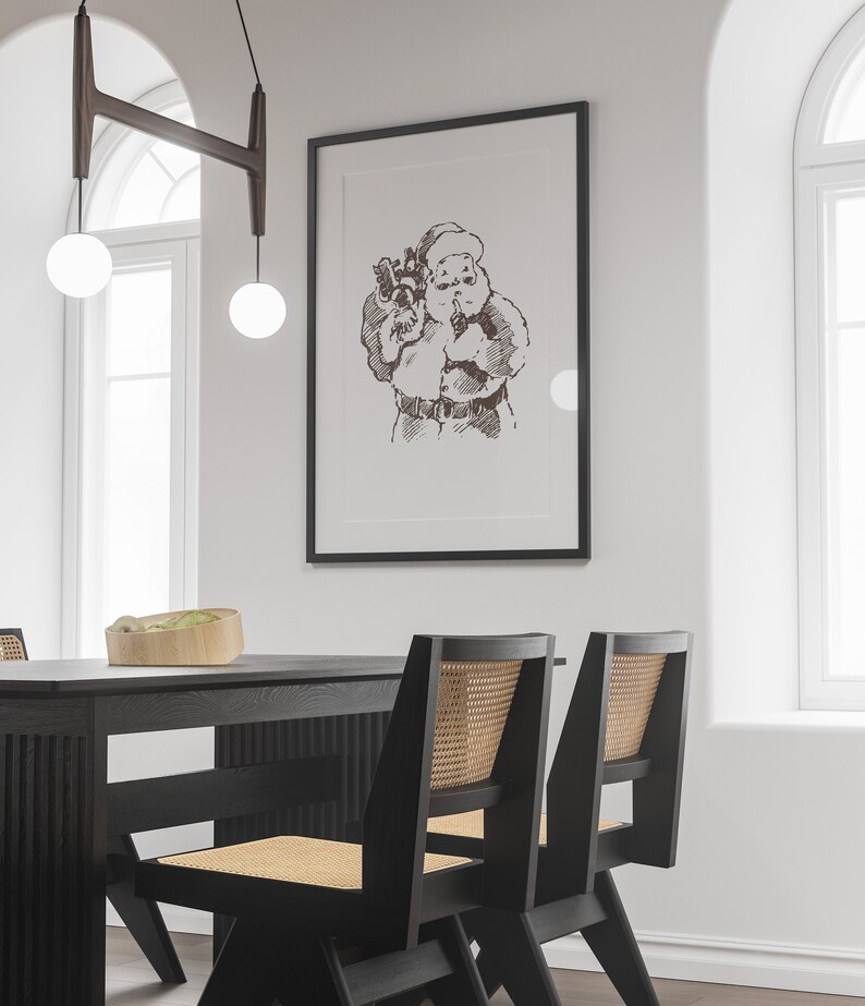May include: A black framed print of a vintage illustration of Santa Claus holding a bag of gifts. The print is hanging on a white wall above a black dining table with two black chairs with woven cane seats.