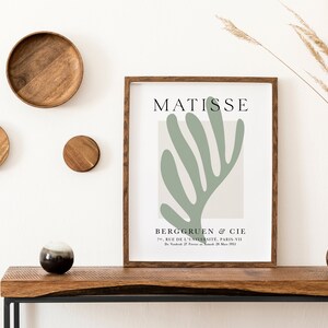 Op de afbeelding: Een ingelijste print van een groen abstract bladontwerp met de tekst "MATISSE" en "BERGGRUEN & CIE" in zwart lettertype. De print staat op een houten tafel met een vaas met droog gras en een kleine bruine keramische vaas.