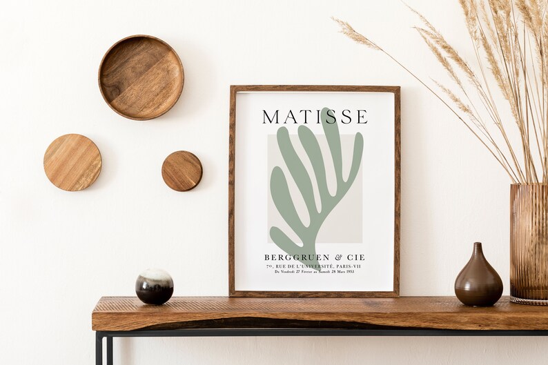 Op de afbeelding: Een ingelijste print van een groen abstract bladontwerp met de tekst "MATISSE" en "BERGGRUEN & CIE" in zwart lettertype. De print staat op een houten tafel met een vaas met droog gras en een kleine bruine keramische vaas.