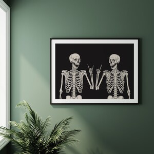 May include: A framed wall art print featuring two off-white skeletons against a solid black background. Both skeletons are depicted from the chest up, each raising one hand to make a rock and roll hand gesture with their fingers pointed upward.