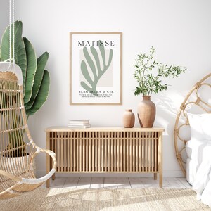 Op de afbeelding: Een minimalistische kunstprint met een groen abstract bladontwerp en de tekst "MATISSE" en "BERGRUEN & CIE" in zwart. De print is ingelijst in een licht houten lijst en hangt aan een witte muur.