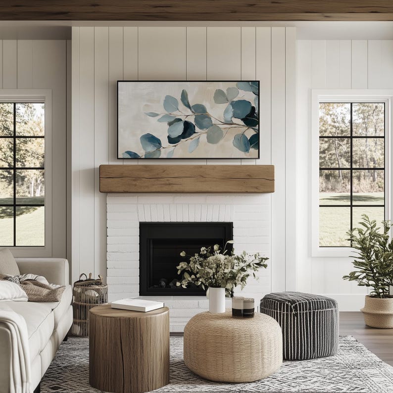 May include: A living room with a white brick fireplace, a wooden mantel, and a framed abstract painting of blue and green leaves. There are two round coffee tables, one with a woven top and one with a wooden top. A white vase with green plants sits on the woven coffee table. A black and white striped ottoman sits in the corner of the room.