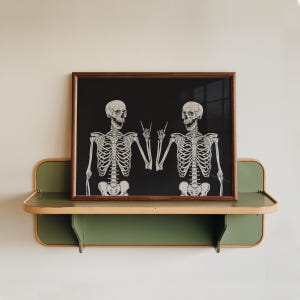 May include: A rectangular wall art print in a dark brown wooden frame featuring a black background with two white human skeletons facing each other, both making a rock and roll hand gesture with their fingers, displayed on a green wall shelf.