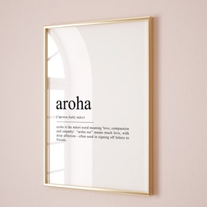 May include: A framed print with a gold frame and a white background. The print features the word "aroha" in black text, with the definition of the word in smaller black text below. The definition explains that "aroha" is a Maori word meaning "love, compassion and empathy".