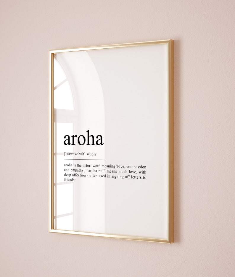 May include: A framed print with a gold frame and a white background. The print features the word "aroha" in black text, with the definition of the word in smaller black text below. The definition explains that "aroha" is a Maori word meaning "love, compassion and empathy".