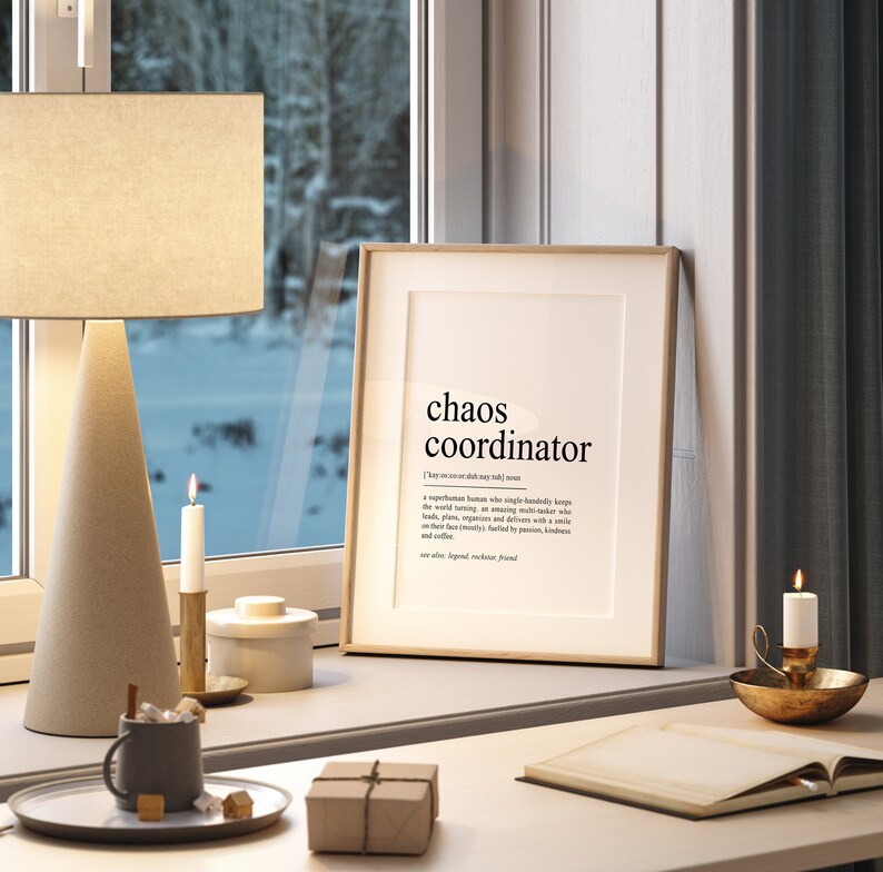 May include: A framed print with a white background and black text that reads "chaos coordinator" and a definition of the term. The print is on a white desk with a brown box, a cup of coffee, and candles.