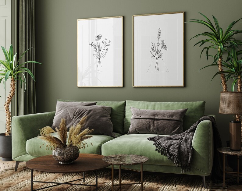 May include: Two framed black and white line drawings of floral bouquets hang on a sage green wall above a green velvet sofa with gray throw pillows and a gray throw blanket.