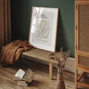 K&ouml;nnte beinhalten: Ein gerahmter Kunstdruck mit einer minimalistischen Linienzeichnung einer Blume in Schwarz auf beigefarbenem Hintergrund. Der Titel des Drucks lautet "Matisse PAPIERS DECOUPES". Der Druck h&auml;ngt an einer gr&uuml;nen Wand &uuml;ber einer Holzbank mit einem geflochtenen Sitz.
