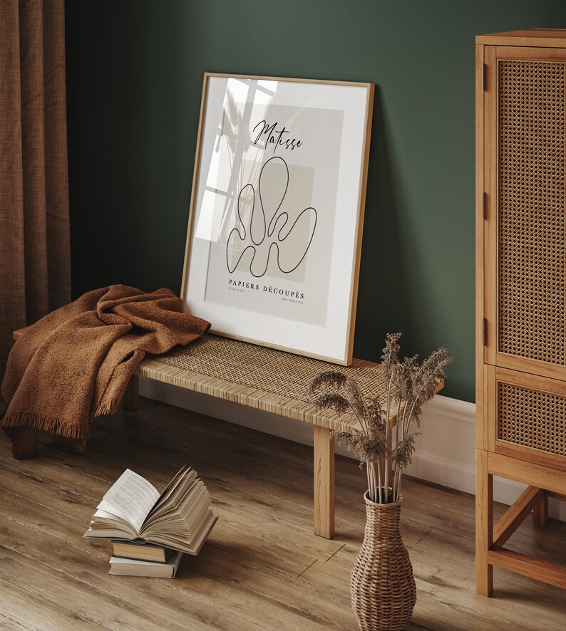 K&ouml;nnte beinhalten: Ein gerahmter Kunstdruck mit einer minimalistischen Linienzeichnung einer Blume in Schwarz auf beigefarbenem Hintergrund. Der Titel des Drucks lautet "Matisse PAPIERS DECOUPES". Der Druck h&auml;ngt an einer gr&uuml;nen Wand &uuml;ber einer Holzbank mit einem geflochtenen Sitz.