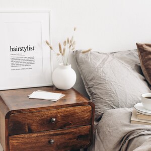 Puede incluir: Una impresi&oacute;n enmarcada en blanco con la palabra "hairstylist" definida. La impresi&oacute;n est&aacute; sobre una mesita de noche de madera con un jarr&oacute;n blanco de flores secas. Una taza de caf&eacute; y una pila de libros est&aacute;n sobre una cama con almohadas marrones y grises.