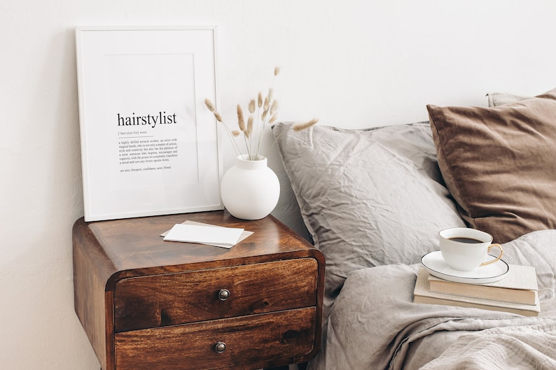 Puede incluir: Una impresi&oacute;n enmarcada en blanco con la palabra "hairstylist" definida. La impresi&oacute;n est&aacute; sobre una mesita de noche de madera con un jarr&oacute;n blanco de flores secas. Una taza de caf&eacute; y una pila de libros est&aacute;n sobre una cama con almohadas marrones y grises.