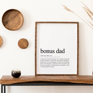 May include: A framed print with a white background and black text that reads "bonus dad" and a definition of the term. The print is hanging on a white wall with wooden decor and a vase of dried grass.