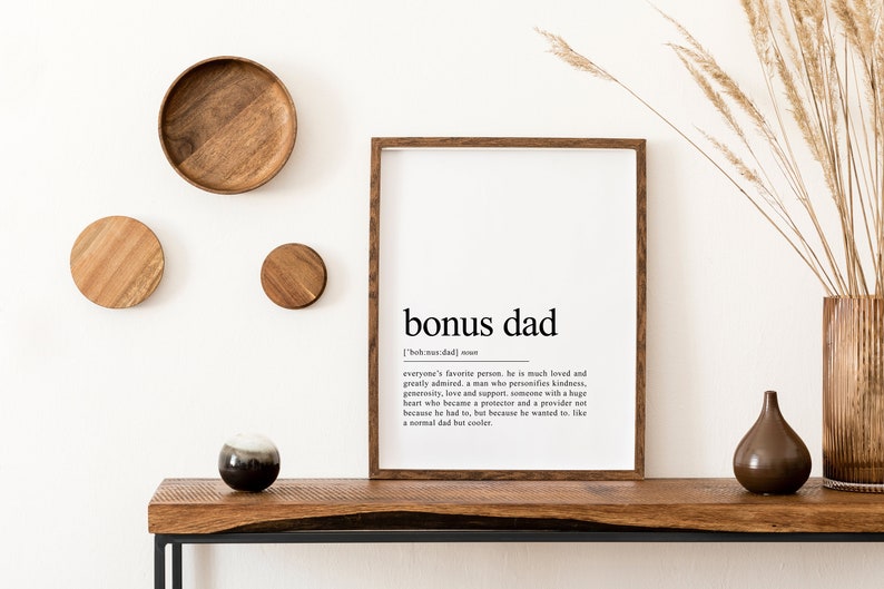 May include: A framed print with a white background and black text that reads "bonus dad" and a definition of the term. The print is hanging on a white wall with wooden decor and a vase of dried grass.