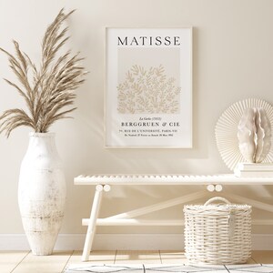 May include: A white framed print of a Matisse painting titled "La Gerbe (1953)" with the text "BERGGRUEN & CIE 70, RUE DE L'UNIVERSIT&Eacute;, PARIS-VII DV 27 25 M 1953". The print is hanging on a white wall above a white bench and a white wicker basket.