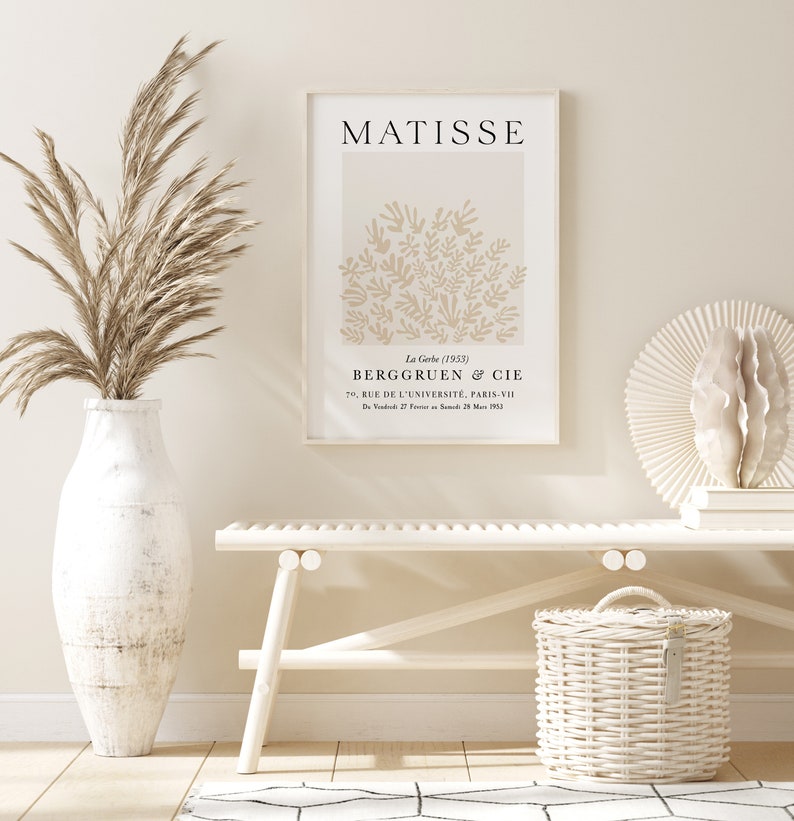 May include: A white framed print of a Matisse painting titled "La Gerbe (1953)" with the text "BERGGRUEN & CIE 70, RUE DE L'UNIVERSIT&Eacute;, PARIS-VII DV 27 25 M 1953". The print is hanging on a white wall above a white bench and a white wicker basket.
