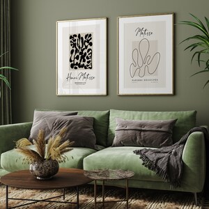 K&ouml;nnte beinhalten: Zwei gerahmte Kunstdrucke mit einem gr&uuml;nen Samtsofa, einer Topfpflanze und einem Couchtisch im Vordergrund. Die Drucke zeigen abstrakte Designs in Schwarzwei&szlig; mit dem Text "Henri Matisse" und "Matisse Papiers Decoupes".