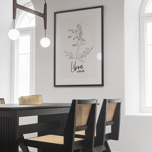 May include: A black and white line drawing of a woman with a scale, surrounded by flowers and leaves. The text "Libra Woman" is written below the drawing. The drawing is framed in a black frame and hanging on a white wall.