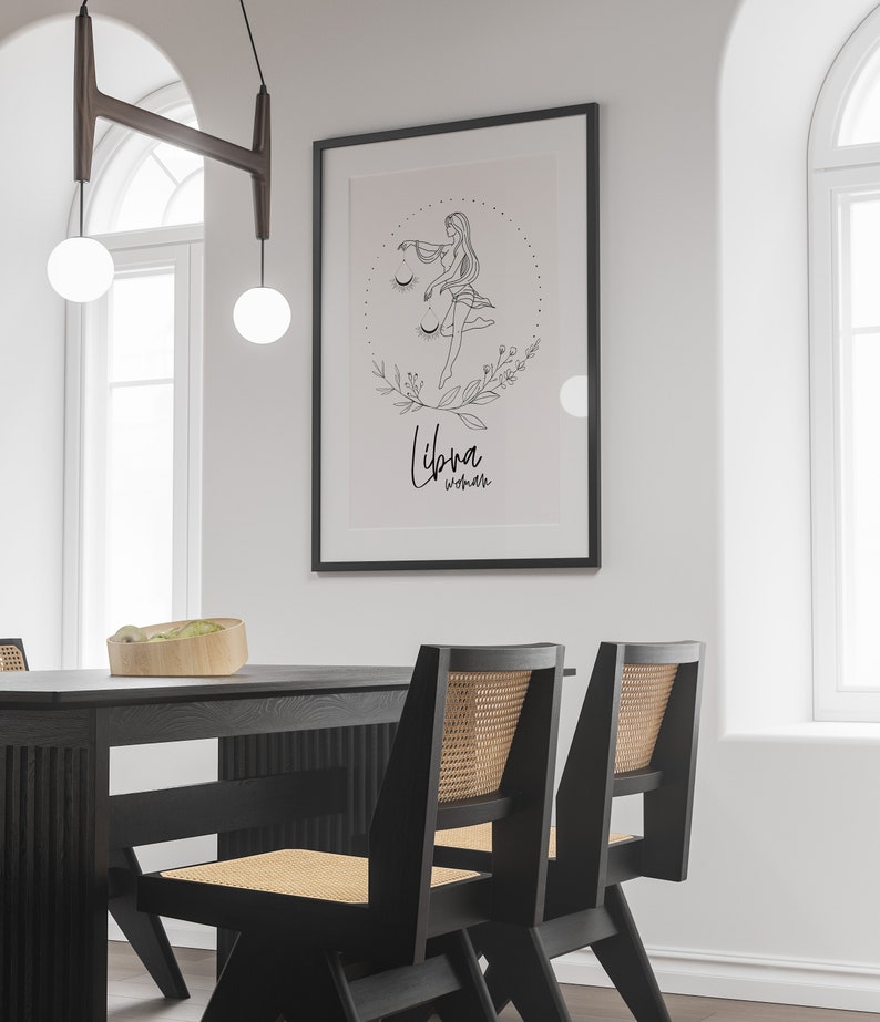 May include: A black and white line drawing of a woman with a scale, surrounded by flowers and leaves. The text "Libra Woman" is written below the drawing. The drawing is framed in a black frame and hanging on a white wall.