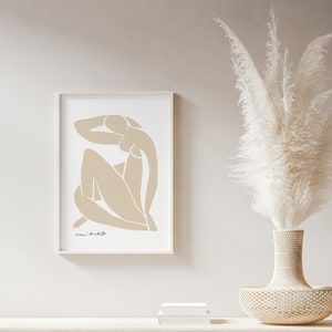 May include: A framed print of a beige abstract line drawing of a person sitting with their arm raised above their head. The print is on a white background and has a white frame. The print is hanging on a light beige wall.